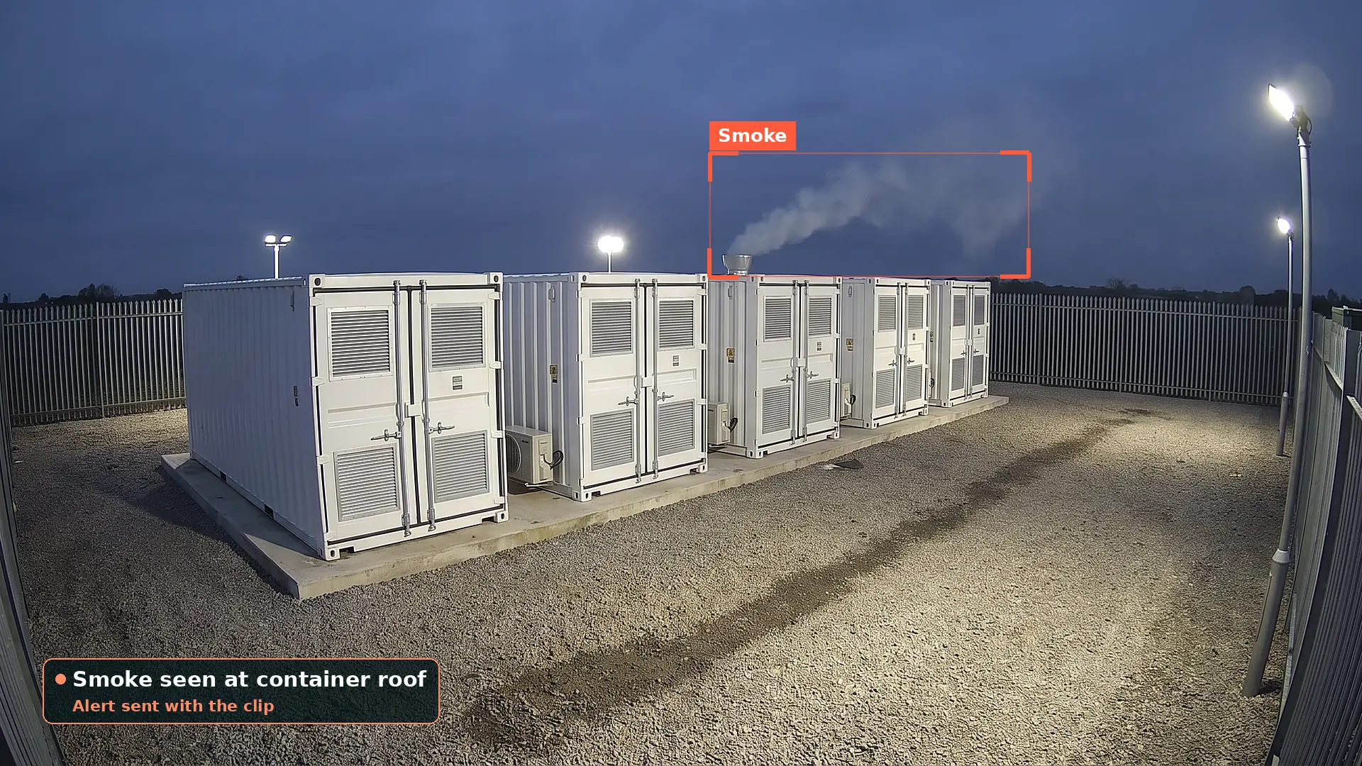 Row of five white battery storage containers on a concrete plinth inside a palisade compound at dusk, with grey smoke rising from a vent stack on one roof, boxed by a red detection box, and an alert card saying the alert was sent with the clip