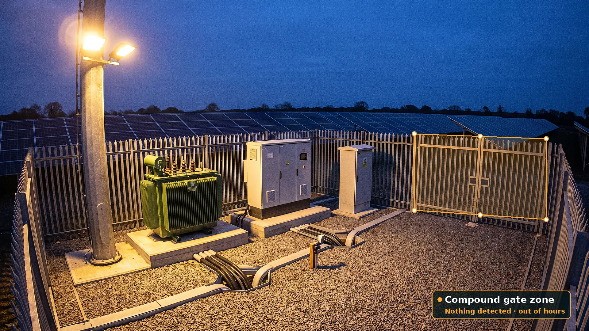 Inverter and transformer station at twilight inside a palisade compound, with a green transformer, a grey inverter cabinet and a kiosk under a floodlight, and an amber polygon zone drawn across the double compound gate