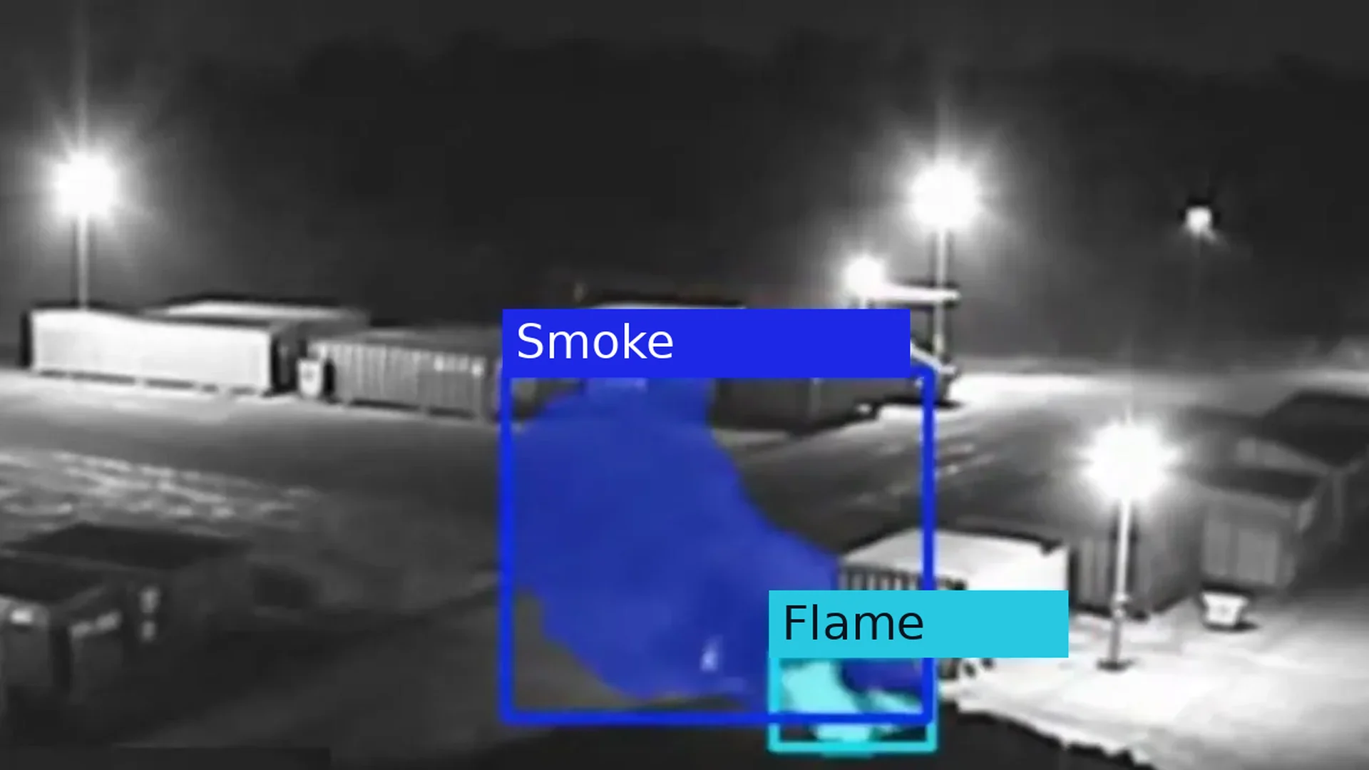 Real night CCTV frame of a waste site container yard under floodlights, with smoke boxed above the end container and flame boxed at its base